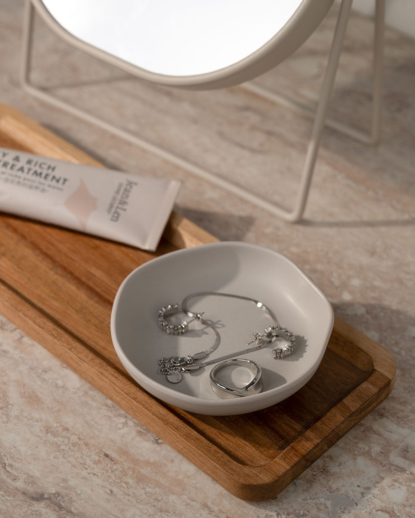 Schmuckschale mit Silberring, Kette und Ohrringen auf einem Holztablett; daneben eine Handcreme und ein Spiegel.