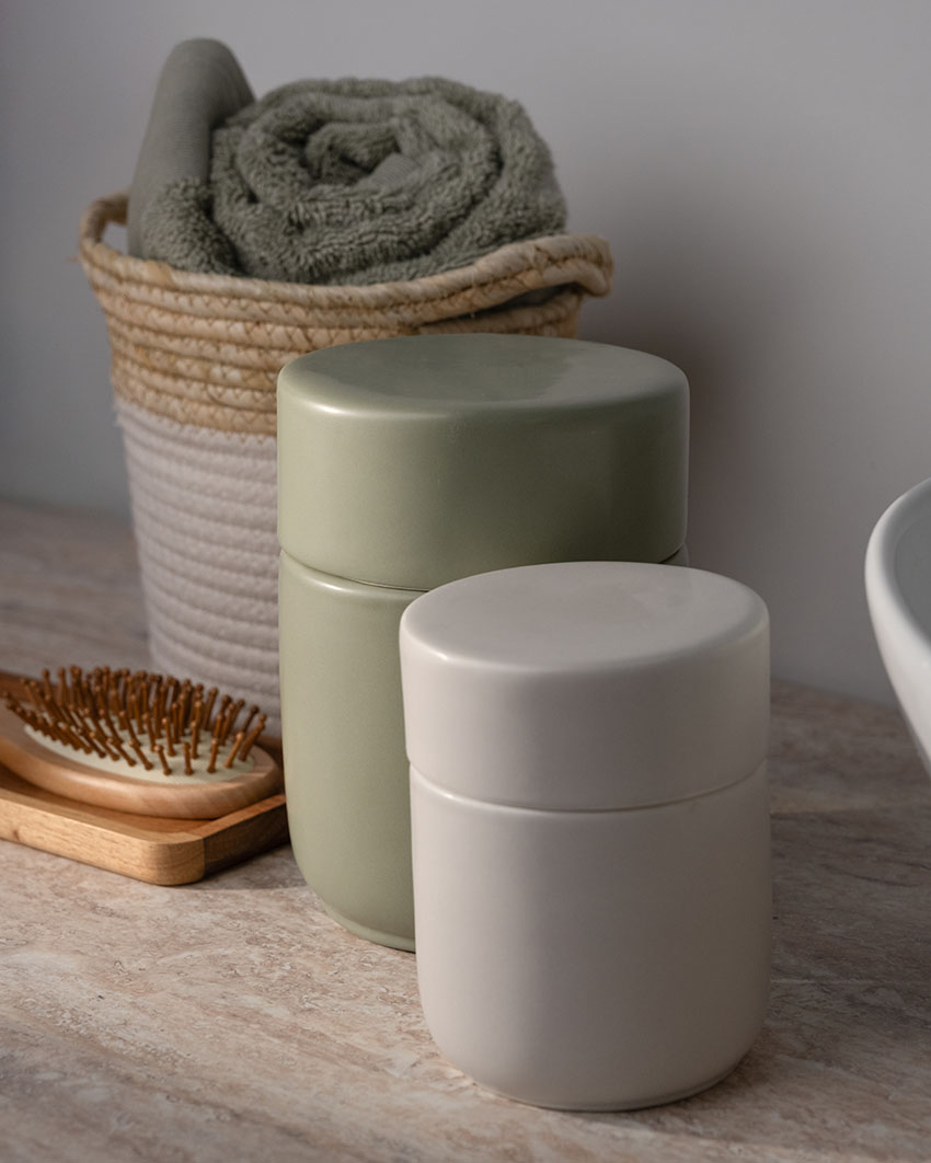 Zwei Aufbewahrungsdosen in Beige und Salbeigrün auf einem Waschtisch, im Hintergrund ein geflochtener Korb mit gerolltem Handtuch.