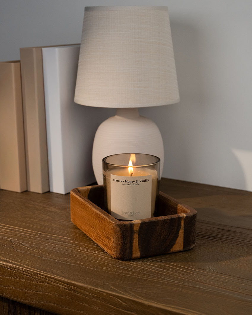 Duftkerze „Manuka Honey & Vanilla“ von Jean&Len brennt in einem Holztablett auf einer Kommode neben einer Tischlampe und Büchern.