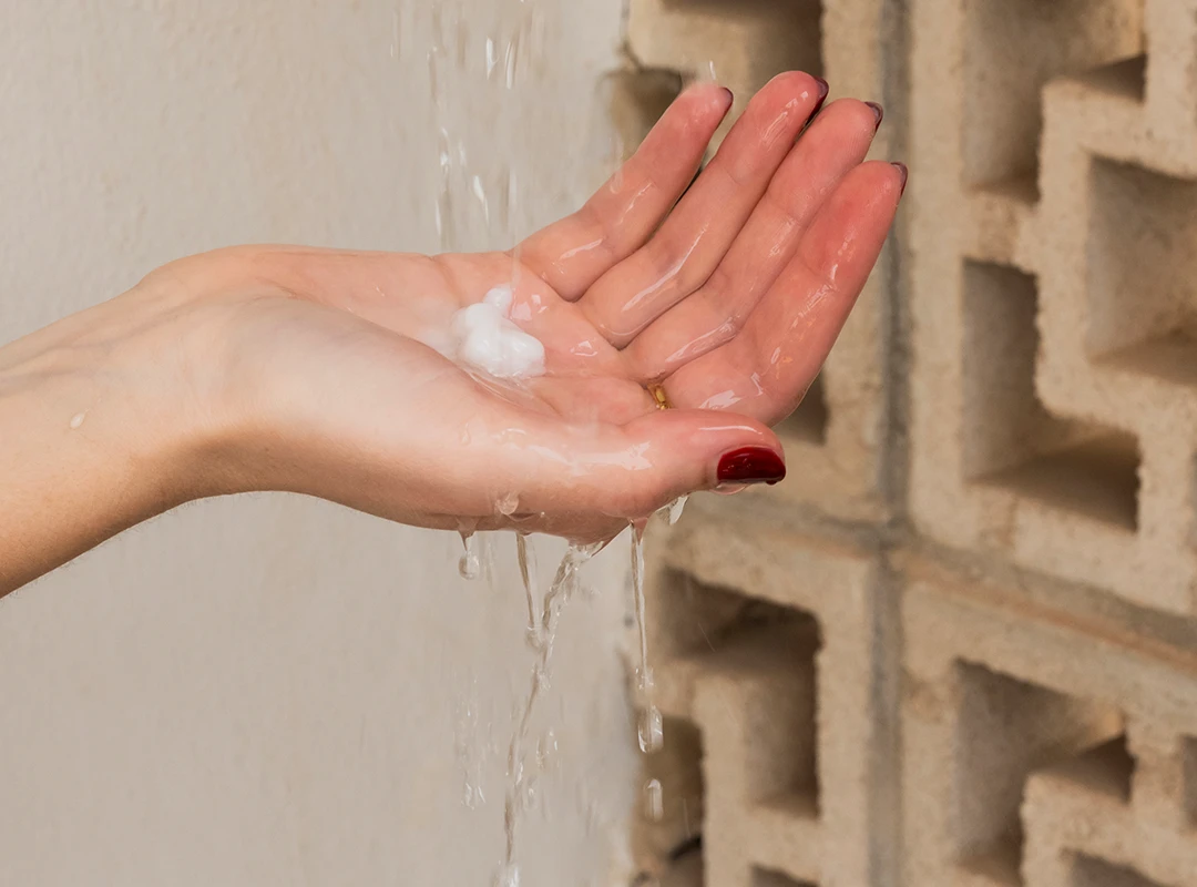 Hand mit Conditioner unter Wasserstrahl vor gemusterter Steinwand – zeigt Anwendung eines Haarpflegeprodukts