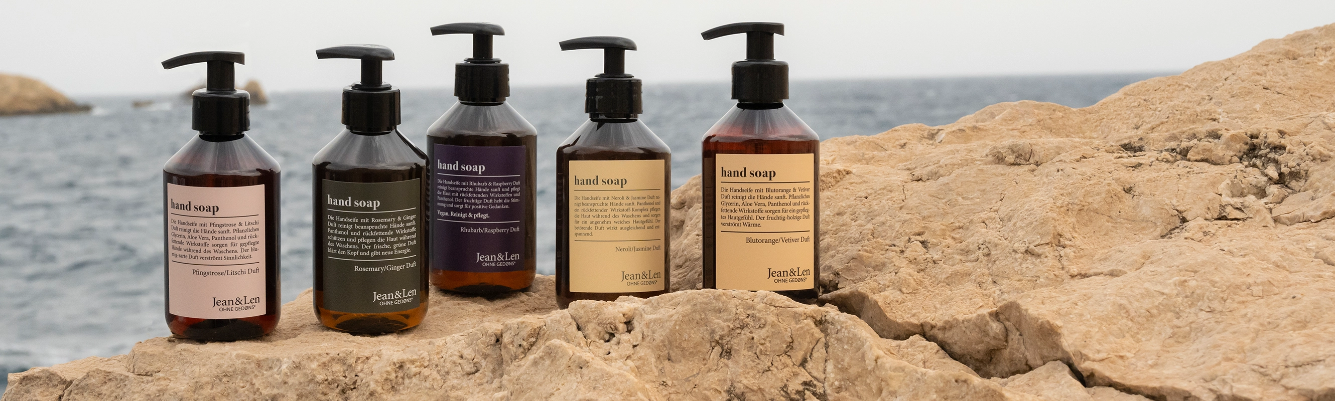 Fünf Jean&Len Handseifen in Glasflaschen auf Felsformation mit Meer im Hintergrund