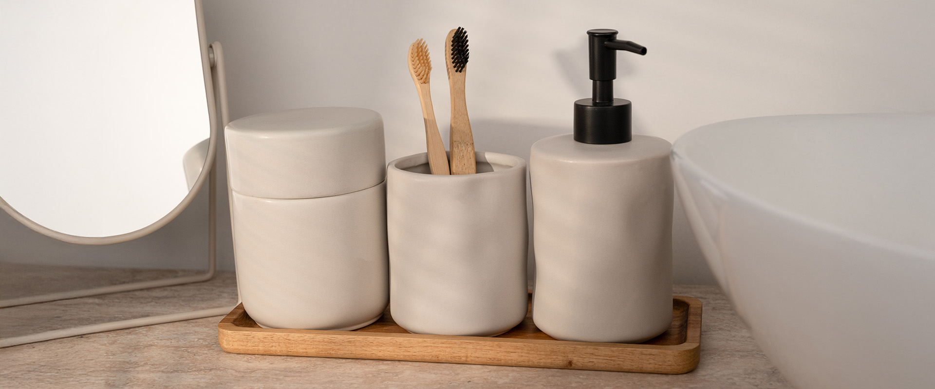 Badezimmer-Set aus Steingut mit Seifenspender, Zahnbürstenhalter und Dose auf einem Holztablett vor einem Spiegel.
